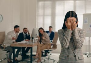 Woman feeling excluded in office while others laugh, symbolizing unfair treatment. Text: "How ‘No Unfair Helping’ Rules Benefit Small Shops | Tech Garcons."
